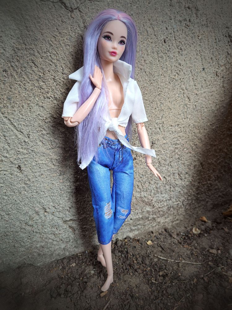 Lavender haired Barbie Doll leaning against a concrete wall. She is wearing jeans and white tied shirt.