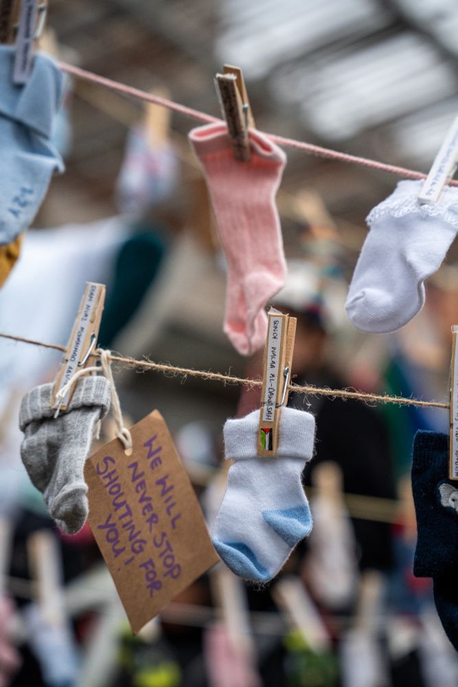 An image of baby socks, pegs hold them onto a line with names of Gazan children murdered in the genocide.
