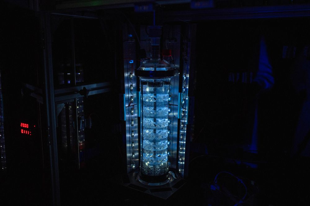 Gerührter Schlaufenphotoreaktor mit eingebauten statischen Mischern (Foto: Elvira Eberhardt / Uni Ulm). Auf dem Bild zu sehen ist eine bläulich leuchtende Apparatur aus Glas und Metall, die in unterschiedliche Reaktionsräume unterteilt ist. 