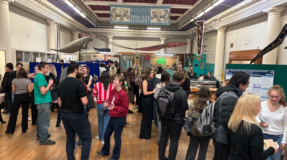 Staff and students mingle and review poster stands in the Graham Kerr Zoology Museum. They are framed from a straight on angle showing the whole room, and under a prominent blue Zoology banner. 