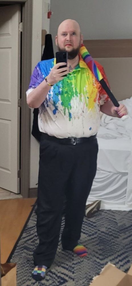 A mirror selfie. I am dressed in a very colorful shirt - rainbow colors melting from the top in a watercolor style down into a white at the bottom. Black pants. One of my socks is a rainbow and the other is checker colors. I have a belt on, a bracelet in the non-binary colors, and I'm holding a baseball bat wrapped in rainbow colors with a black handle. My eyes look a little crooked because of bruising from my new piercing but I'm definitely smiling.