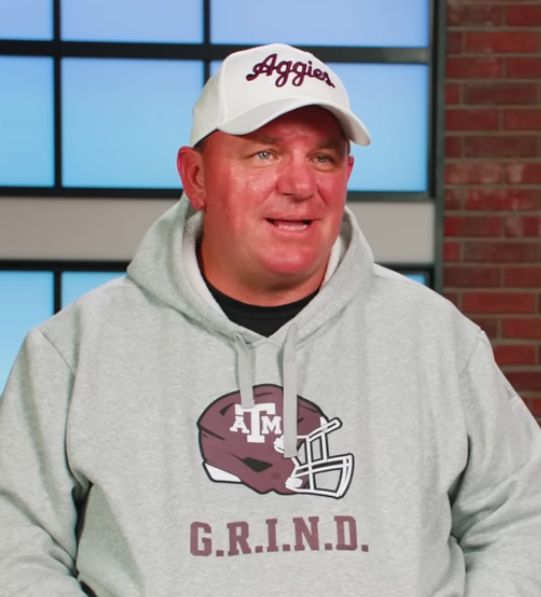 Mike Elko in a white hat and gray GRIND hoodie looking extremely sunburnt