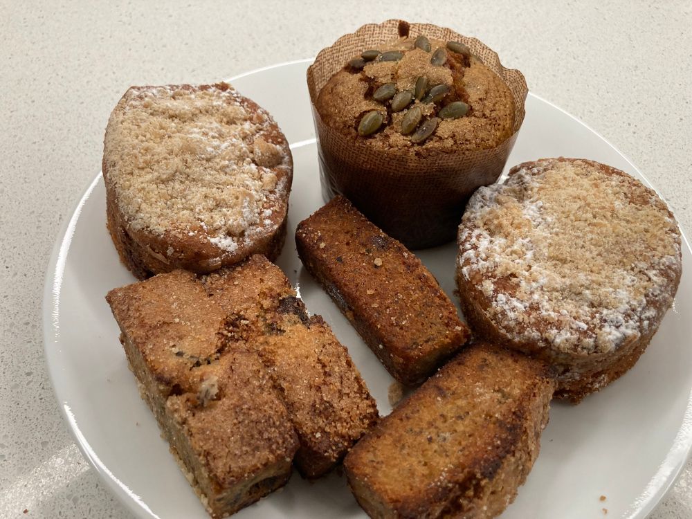 White plate with a pumpkin muffin topped with seeds, two coffee cakes, and four slices of cherry bread
