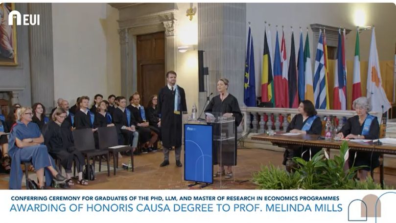A woman in stands at a podium to address an audience. There is a line of international flags in the background and all the attendees of the event are wearing academic address. A caption below the image reads 'Awarding of honoris causa degree to Prof. Melinda Mills'.