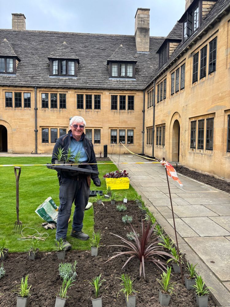 Walter Sawyer stands behind one of the beds in the Upper Quad of Nuffield College, holding a tray of plants. In front of him are various bedding plants laid out in small pots to be planted.