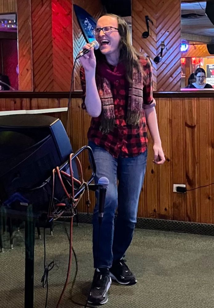Me in a red and black flannel shirt, pink scarf and jeans, singing “You Oughta Know” at karaoke 