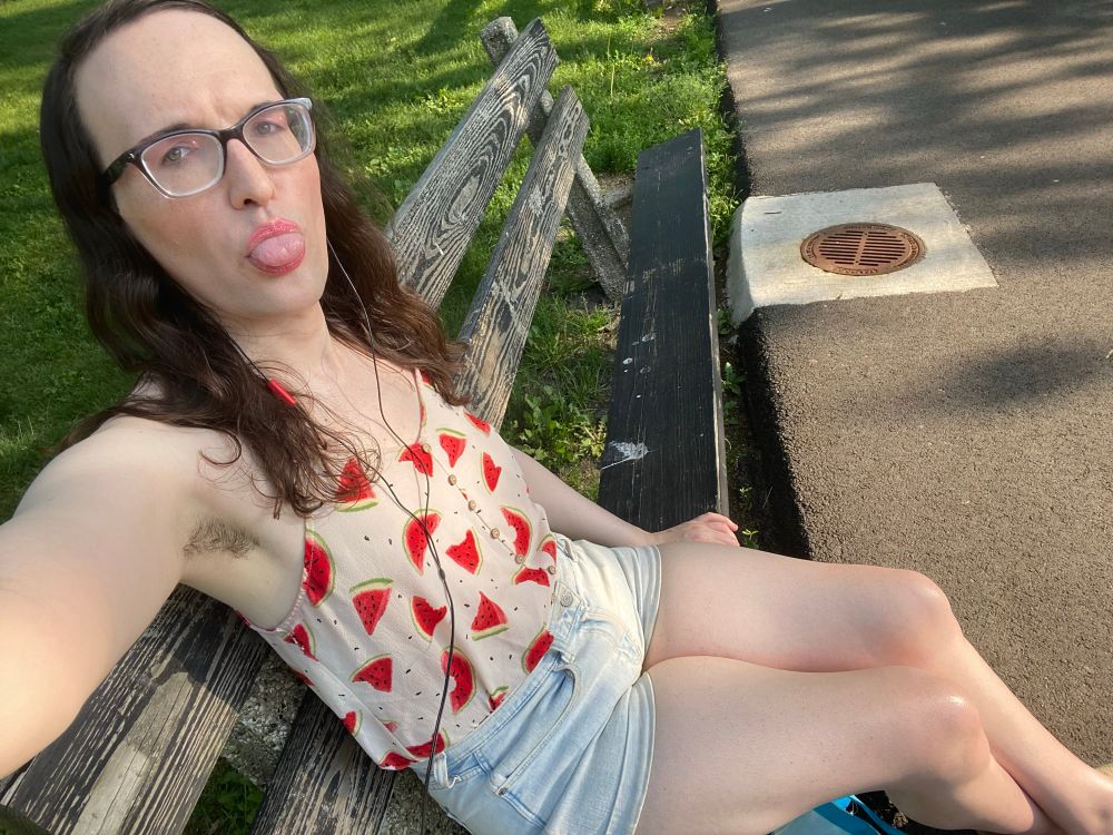 Me on a park bench, sticking my tongue out, wearing faded light blue jean shorts and a white top with watermelon slices on it