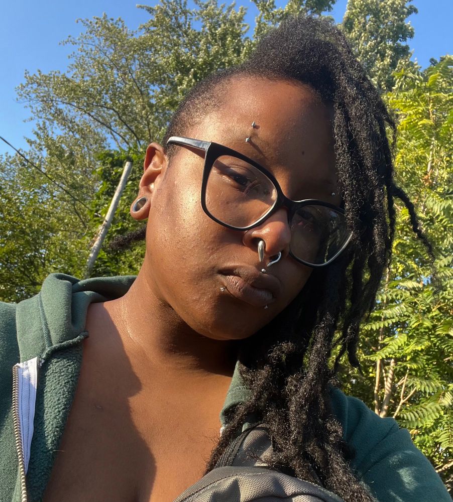 A selfie of salem in a green zip up hoodie wearing black glasses outside in front of green trees