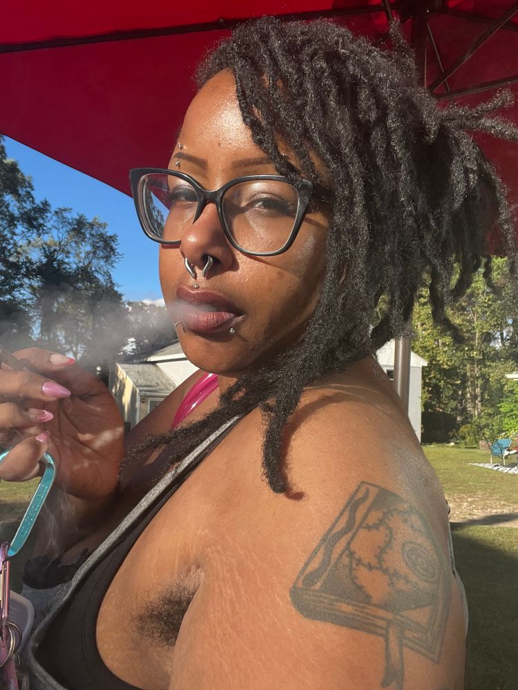 Selfie of a fat Black intersextfemmeangelbear with long locs up in a bun with bangs, wearing a grey tanktop and black glasses, holding a large blue carabiner on her little finger and a joint between her pointer and middle, looking at the camera while exhaling a puff of smoke
