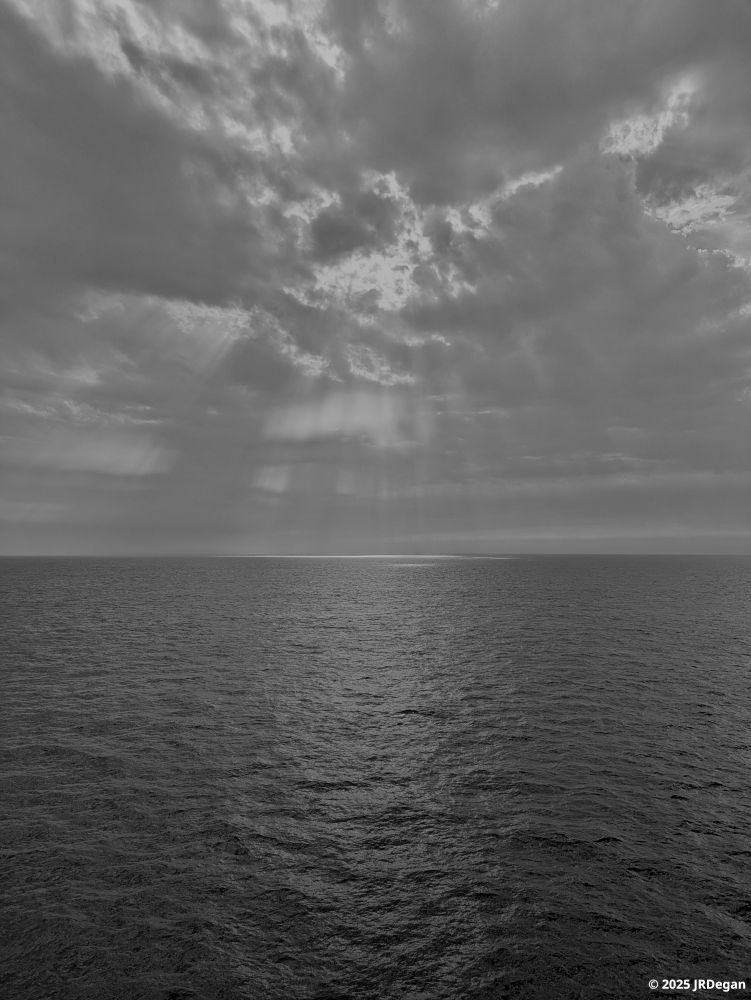 Black and white seascape.  Rays of sunlight streaming through the clouds over a sea that looks like molten lead.