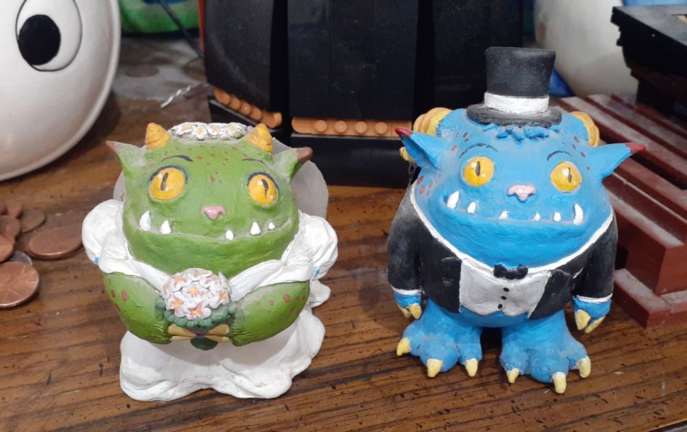 A green monster with yellow googly eyes and little yellow horns, wearing a white wedding dress and carrying a bouquet of flowers, standing next to a blue monster with yellow googly eyes and big yellow horns in a tuxedo and a top hat. 

They are married. Maybe even to each other, maybe. 