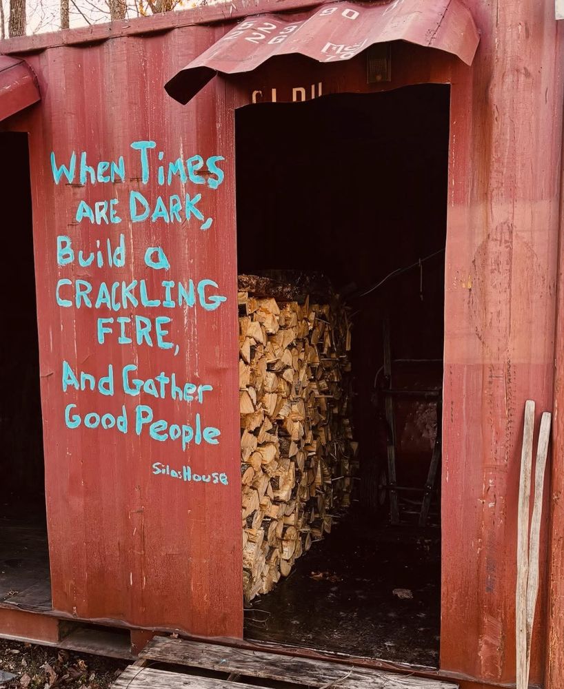 A poem by Silas House painted on the side of a shed of firewood. Poem reads "When times are dark, build a crackling fire, and gather good people."