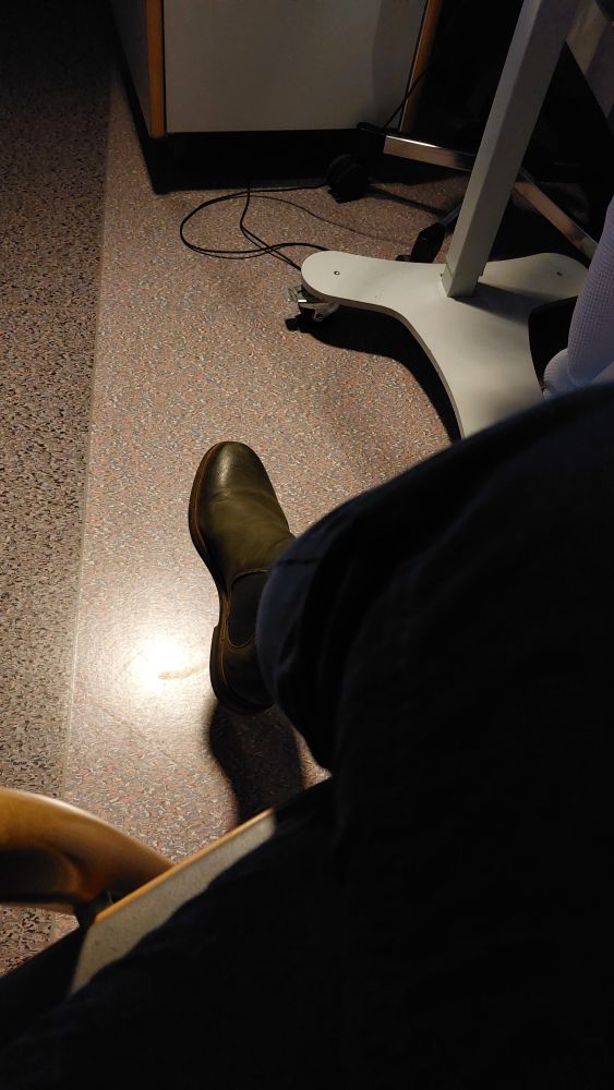 Person mit dunkler Cordhose und dunkelgrünem Schuh sitzt am Spitalbett. Nur Bein, Schuh und Boden sind sichtbar. 