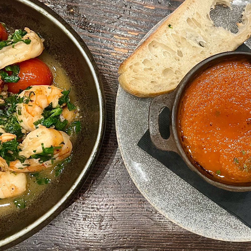 A picture of a plate of shrimps and tomatoes with cilantro on the left and a bowl of tomato soup and a slice of baguette to its right.