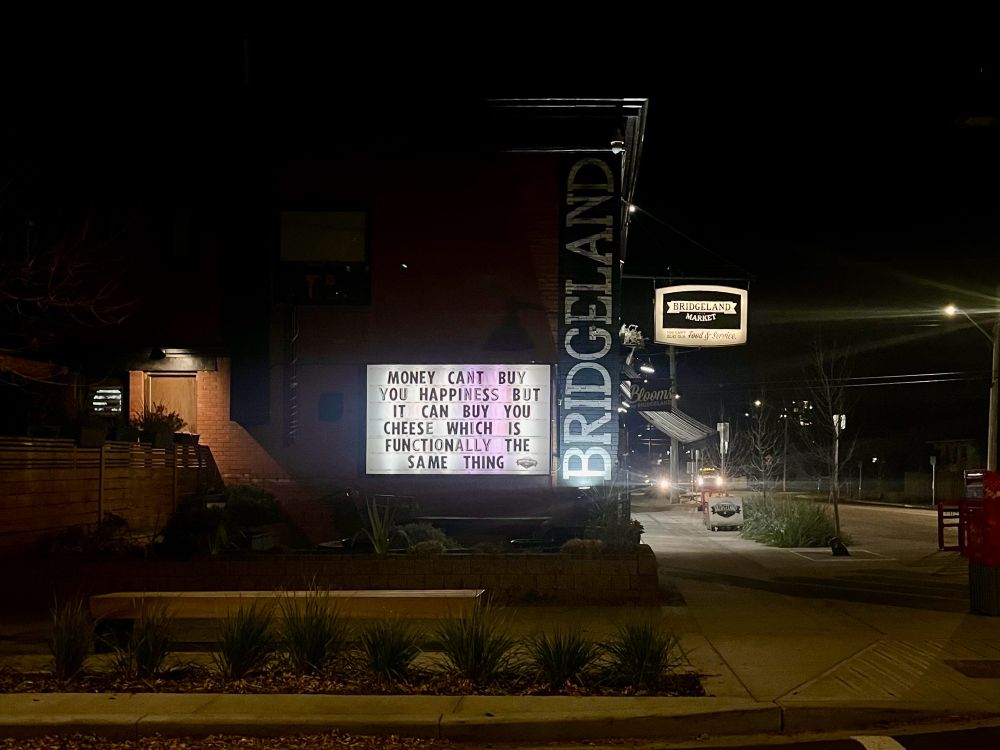 Market sign that say “Money can’t buy happiness but it can buy you cheese which is functionally the same thing”.