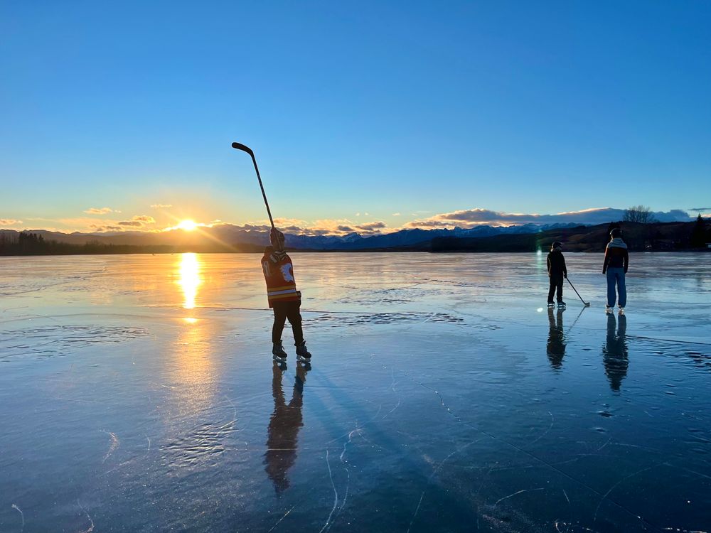 Sun sets down the Rocky Mountains with one person holding their hockey stick up and two others chat in the background.