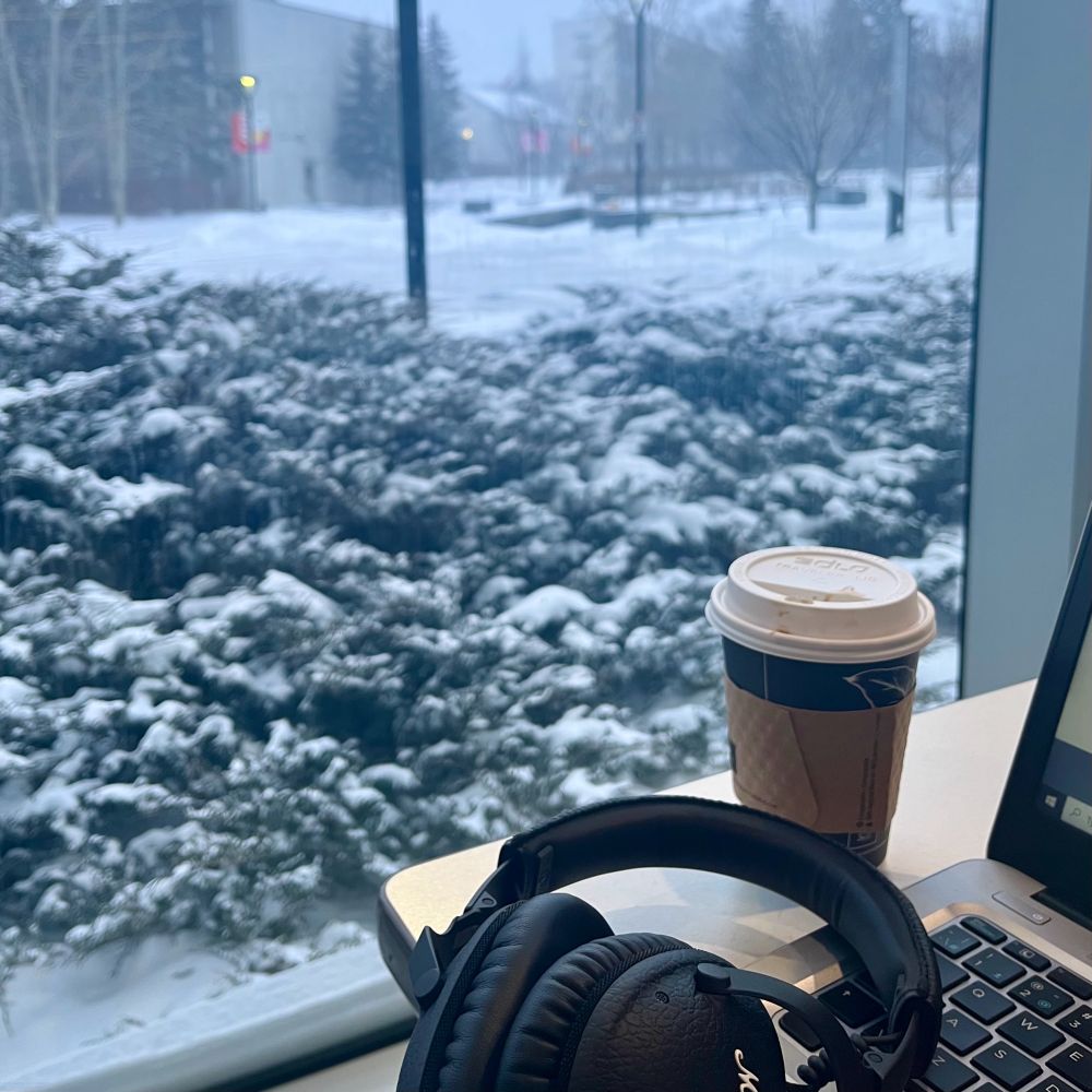 A cup of coffee, headphones, and a laptop are set on a table beside a glass window that shows a snowy scenery including snowed on bushes and pavement.
