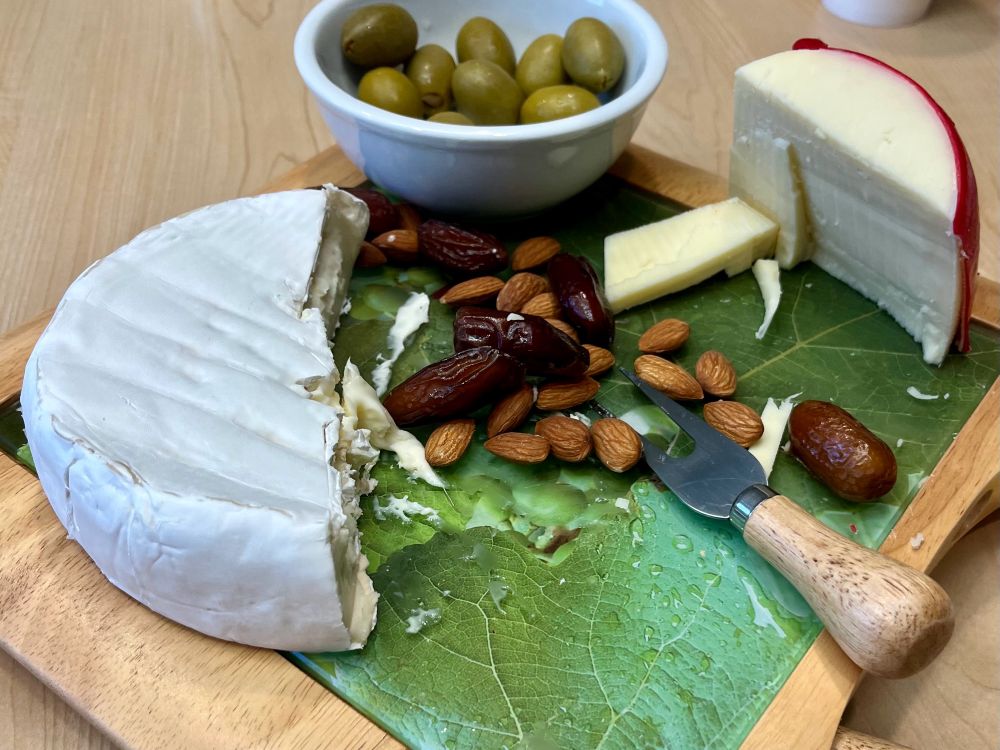 Small cheese board with dates and walnuts spread across it, half of a whole Brie cheese, a bowl of green olives, and Edam cheese.