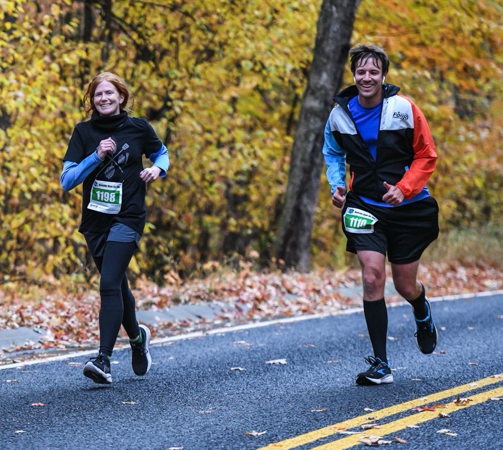 A man and a woman running side by side during a 26km road race. Fallen leaves litter the ground, and the background includes trees with yellow leaves.