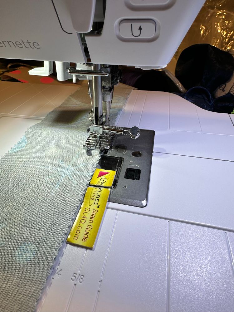 A sewing machine with a table attached and fabric in the process of being sewn. A yellow sticker helps guide it where it needs to go. 