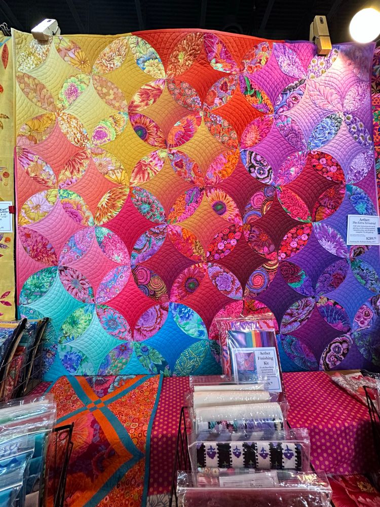 Picture of a quilt hanging on a show wall. It is made up of colorful circles that have solid(ish) color diamonds in the center and florals around the outside of those diamonds to make the circle complete. The colors fade in and out from yellow to red to pink to blue to purple. 