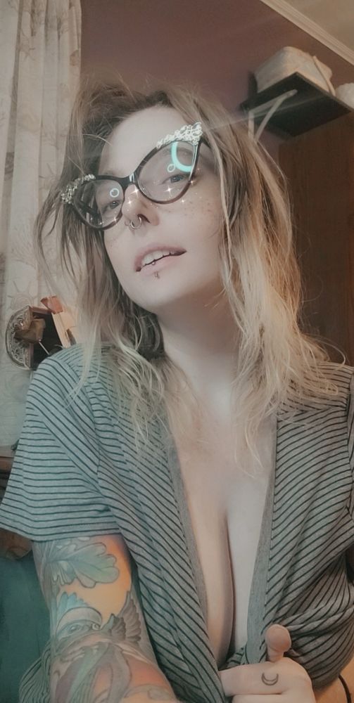 Blonde haired girl with glasses and facial piercings tugging her grey and black striped shirt down in the front to show some of her cleavage 