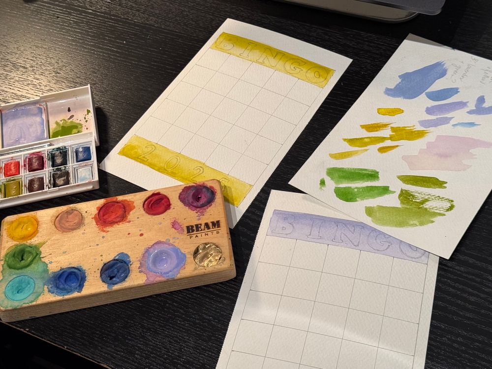 A desktop with some watercolour paints and paint swatched on paper in shades of olive greens, lavender, chartreuse, and periwinkle 