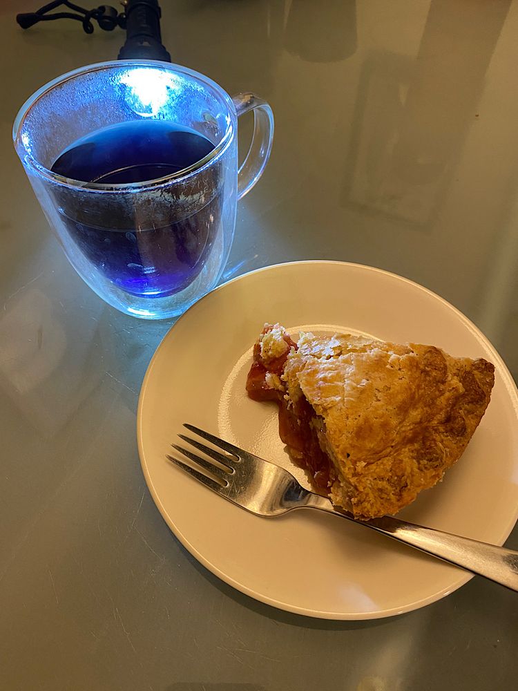 Photo of a slice of strawberry rhubarb pie on a plate and a glass of blue pea flower tea. A flashlight shines behind the tea to better show its blue colouration. 