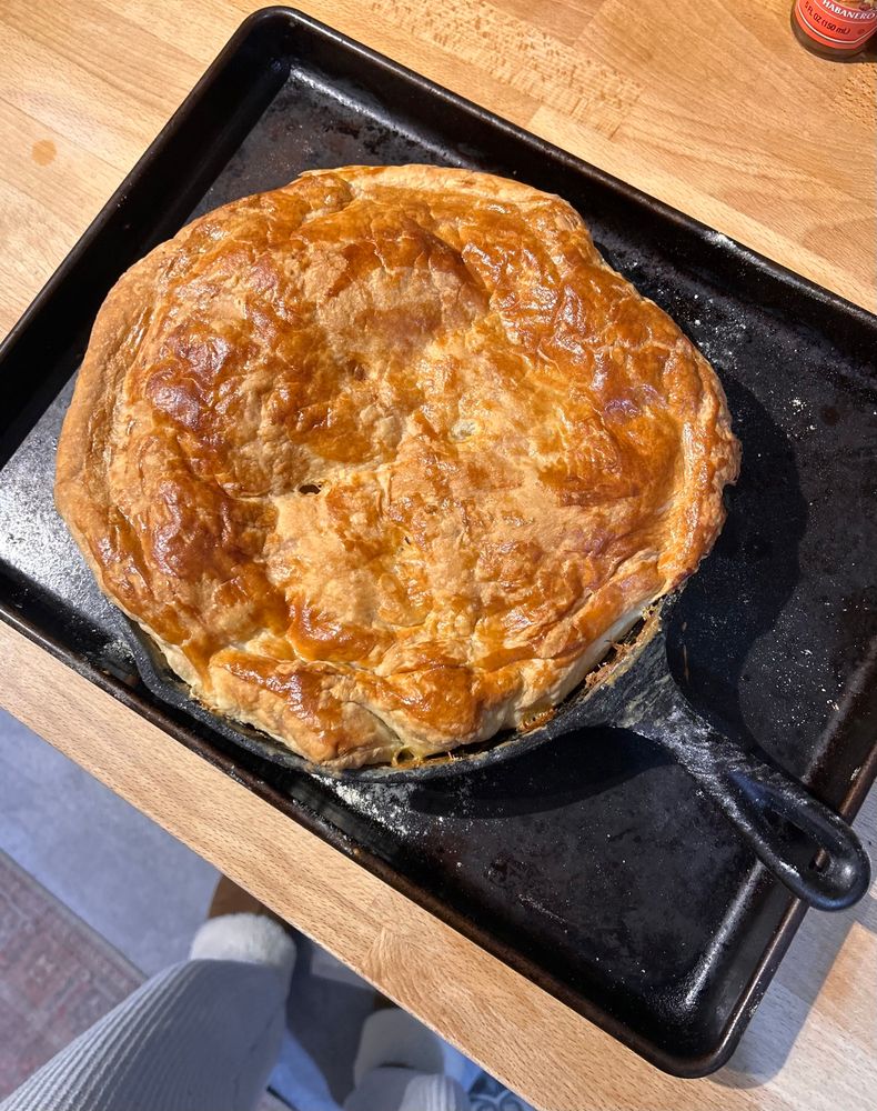 A Pot Pie in a cast iron skillet with a golden brown pastry topper resting on a well-loved sheet pan as it cools on a butcher block countertop 