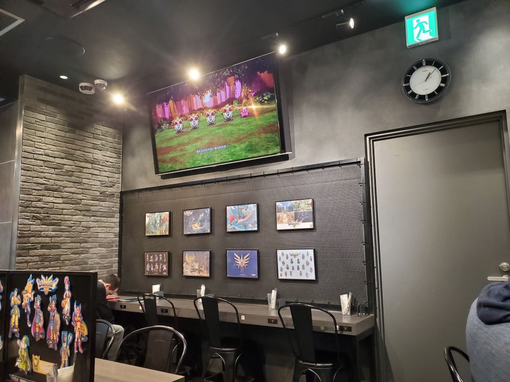 a typical wall decor in the square enix Cafe, with a handful of dragon quest posters and a television showing gameplay of one of the games