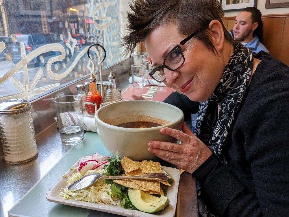 A white lady with a mohawk sitting at a diner counter smiles impishly, caressing a huge white bowl with dark red soup in it. There is a plate with sliced radishes, cabbage, avocado, corn chips, and cilantro. 