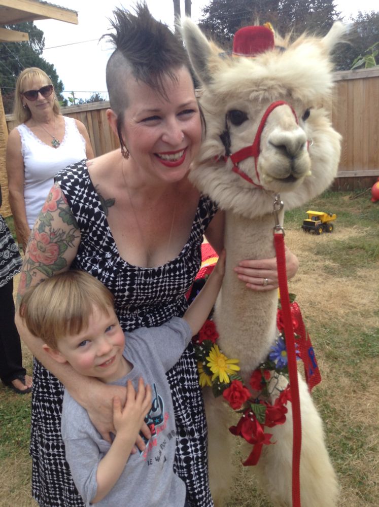 A buff alpaca with a tiny red fez on being hugged by a white woman with a mohawk and a little white boy and everyone is smiling. 
