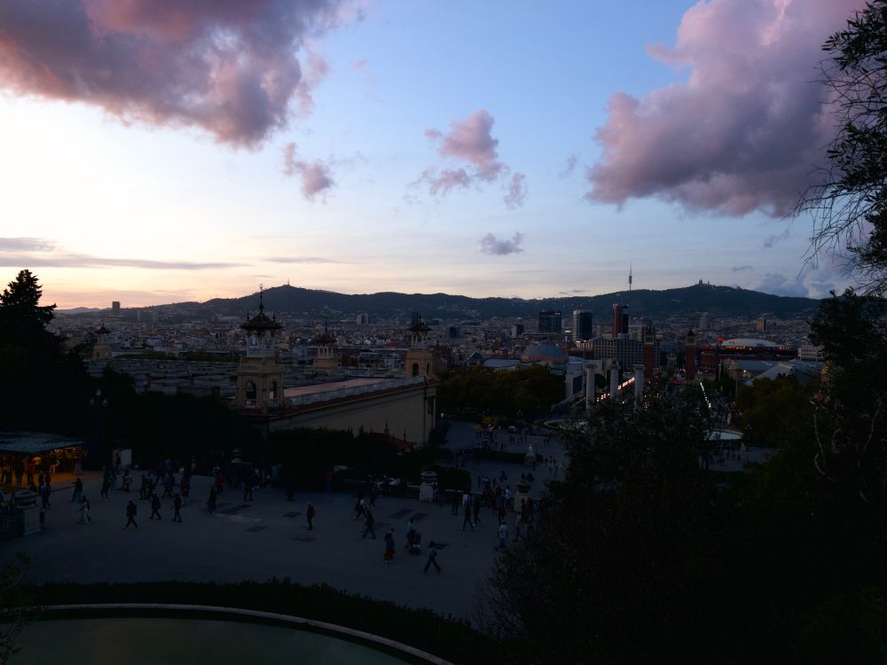 View of Barcelona from a high place showing terraced open spaces going down level by level to the city, with people milling all about. Fountains and cafes visible.