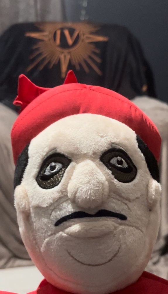 The face of a plushie based on Cardinal Copia from the band Ghost, commonly known as “Plushia”. He is extremely ugly. The background features a black and Ross gold Papa Emeritus IV blanket on the back of an armchair