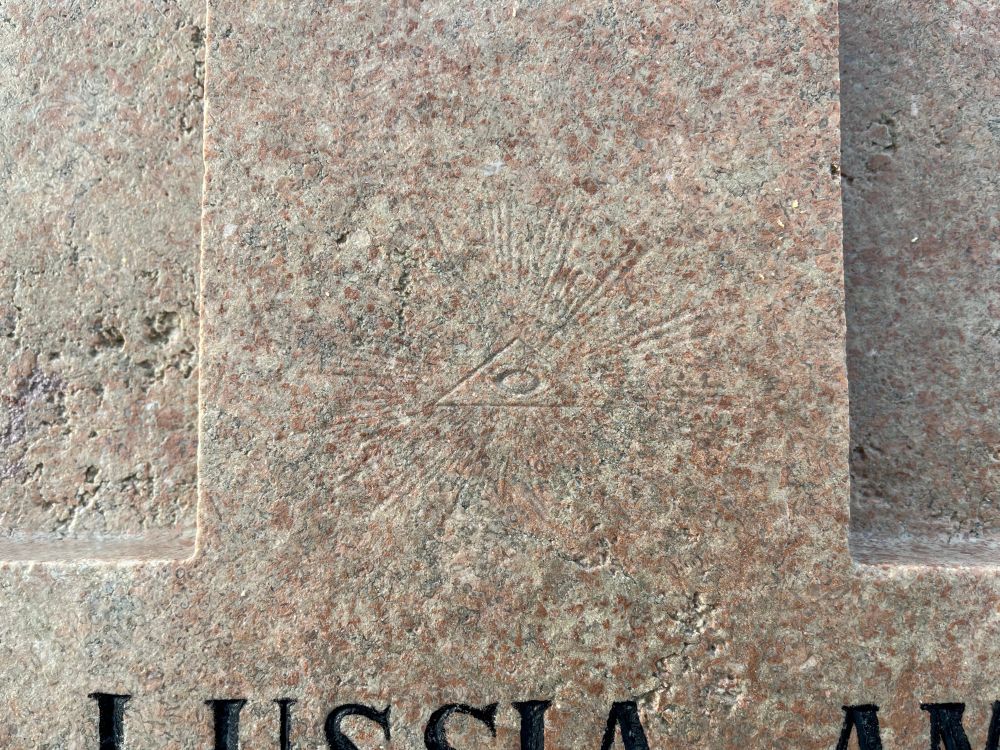 Gravvård i grårosa kalksten med en relief med strålar ut från en platt triangel med ett öga i