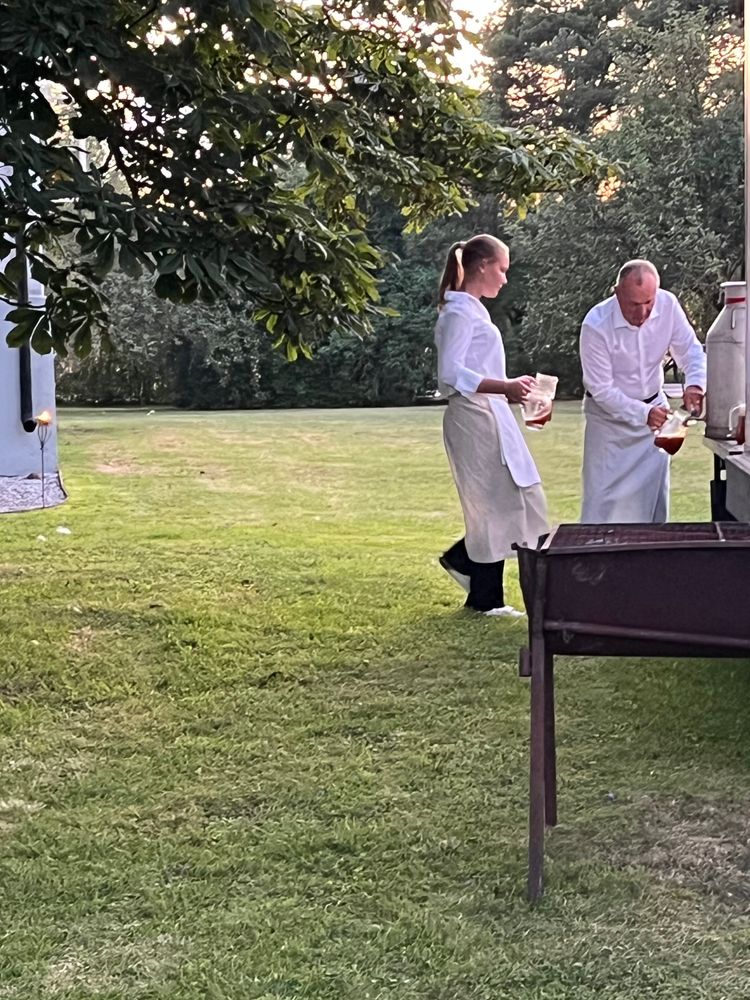 En man i förkläde kranar upp en bärnsstensfärgad vätska från en 40-liters mjölkkanna i aluminium ute på en gräsmatta vid ett bord. Bredvid hon kommer en kvinna i förkläde med två tomma kannor för att fylla på.