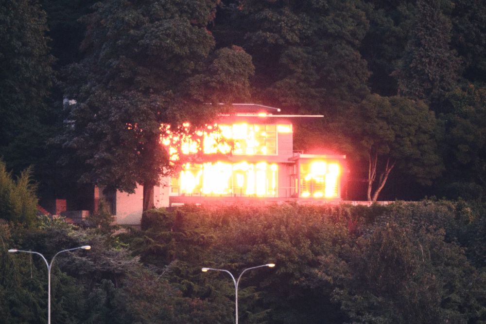 A hyperzoom shot of the lit-up house from the last photo. Barely larger than a speck in that wide shot, the house now takes up probably half of the frame. It legitimately looks like it's on fire, the house's many large windows are producing dazzling reflections of the setting sun. The house is surrounded by green trees, and a couple tall highway lamp posts rise up from beyond the bottom of the frame.