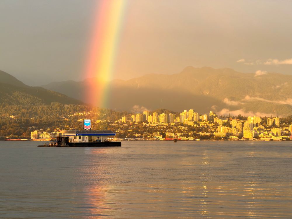 A rainbow ends at the surface right in line with the floating Chevron seaplane fuel station in the Vancouver harbor. Amber sunset / post-storm light washes over North Vancouver and the mountains.