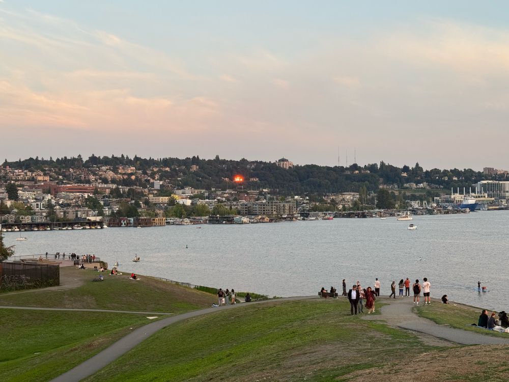 A relatively wide angle photo of the Eastlake area in Seattle as viewed from Gas Works Park. Foreground is a grass hill with a few paved paths and plenty of people. Behind that is the water of Lake Union, then Eastlake and northern Capitol Hill rise out of the water. The hill is probably half trees and half buildings, and one lone building in the center of the hill looks like it's on fire from how it's catching the sun. The sky is partly blue and partly cloudy, the clouds are a warm orange grey.