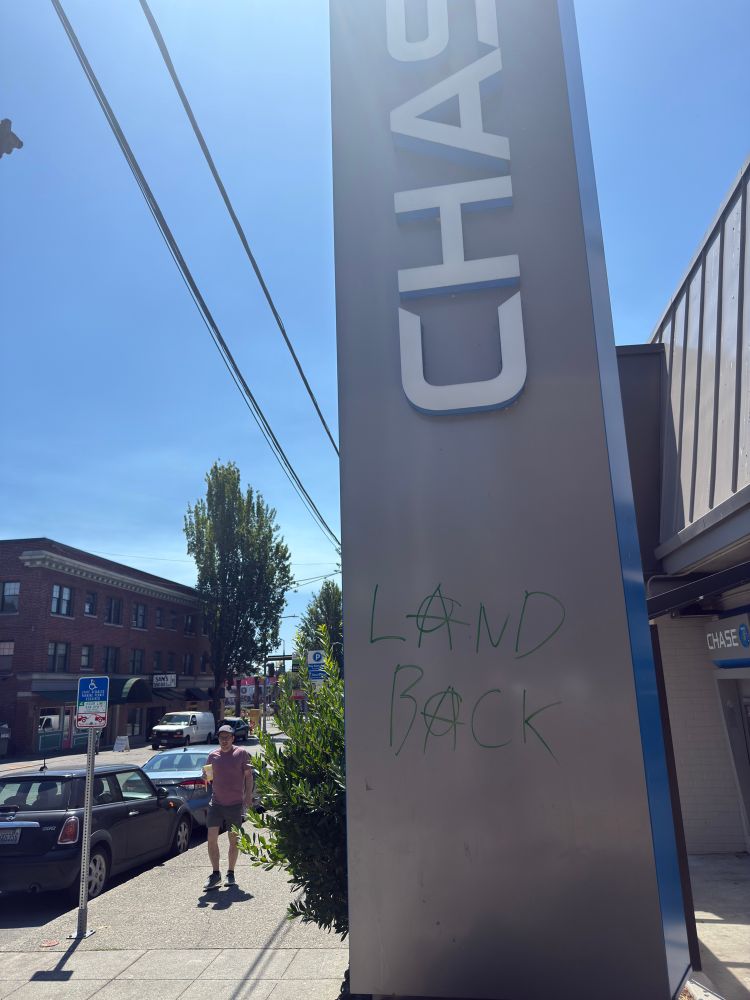 A vertical chase bank sign against a sidewalk and a blue sky. The words "LAND BACK" are written in green ink under the "CHASE" lettering. The "A"s in "LAND BACK" are anarchist circle-As.