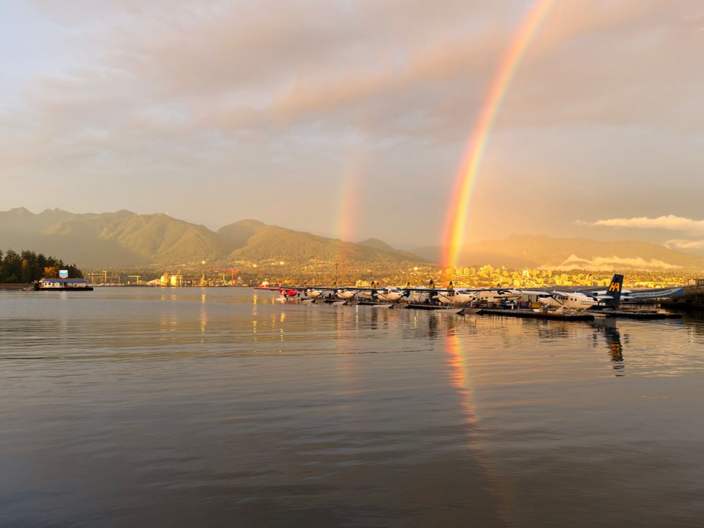 A double rainbow over a row of seaplanes in Vancouver Harbor, flanked by North Vancouver and the mountains. The Chevron seaplane fuel station is floating off to the left behind the seaplanes.