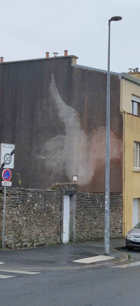 Le mur latéral, sans fenêtres, en béton, d'une maison est mouillé et donc foncé, sauf une forme floue prenant un tiers du mur.