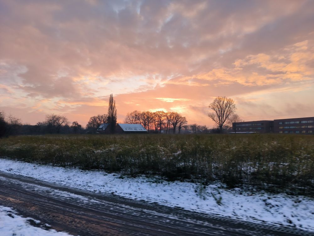 Sonnenuntergang, vorne ein Fahrrad- und Fußweg, dahinter ein Feld und Häuser/Büros. Vereinzelt Bäume und alles ein bisschen Schneebedeckt, was ein guter Kontrast zum orangenen Himmel ist.