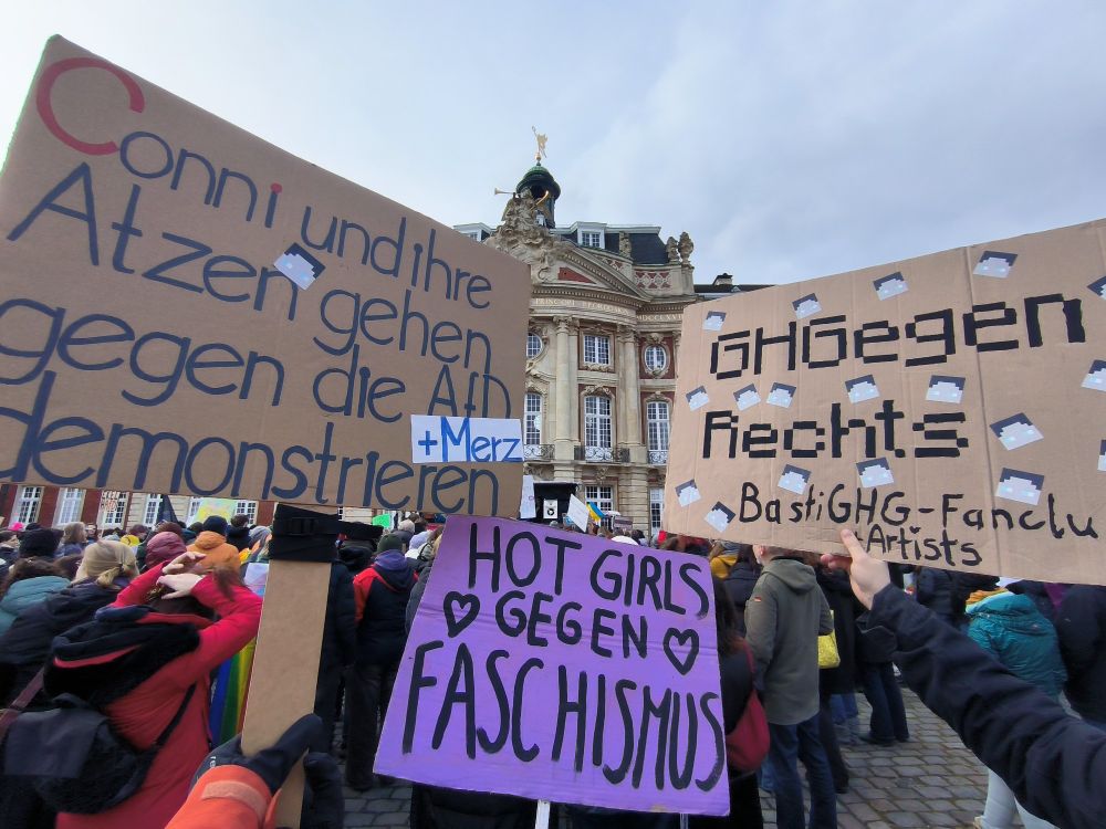 Demo auf dem Schlossplatz, Blick Richtung Schloss. Drei Plakat, die die Sicht beschränken:
1. Conni und ihre Atzen gehen gegen die AfD (+ Merz) demonstrieren 
2. Hot Girls gegen Faschismus
3. GHGegen Rechts (BastiGHG-Fanclub + Artists)