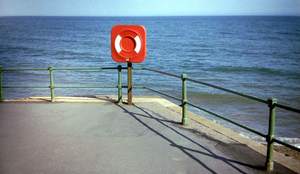 A red and white lifebuoy on a stand. Green railings fringe a promenade. Beyond, a calm sea.