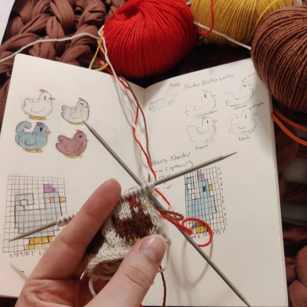 Hand holding a small knit swatch of a brown chicken. In the background is a sketchbook with drawings of stardew valley chickens and three small skeins of yarn in red, yellow, and brown.