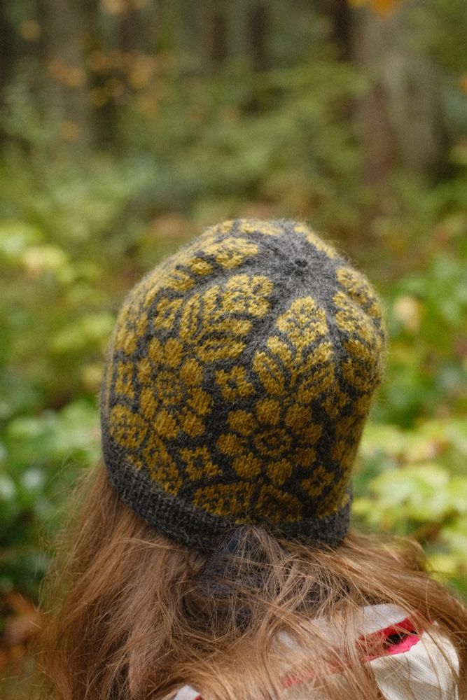 Wearing my newly knit Alpine Bloom Hat out on a hike. The hat is dark grey with a light green floral pattern. Behind me are green and yellow leaves and ferns.