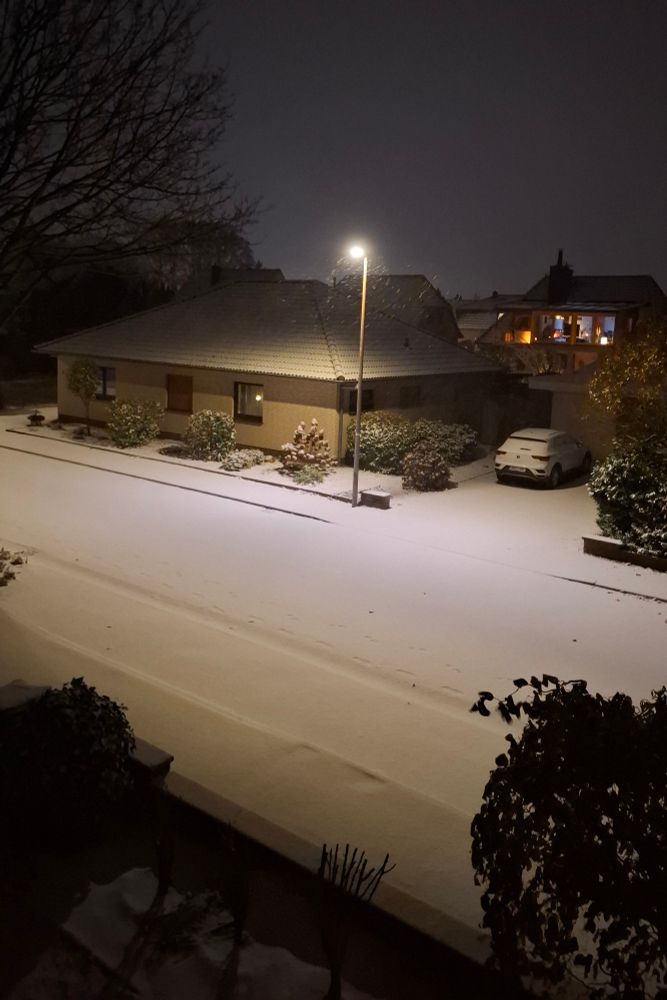Ein nächtliches Winterfoto zeigt eine ruhige, schneebedeckte Wohnstraße. Eine Straßenlaterne beleuchtet den frisch gefallenen Schnee, der Fahrbahn, Gehweg und die Dächer der Häuser bedeckt. Im Vordergrund sind verschneite Büsche und eine Mauer zu sehen. Auf der rechten Seite steht ein Auto in einer Einfahrt, ebenfalls von Schnee überzogen. Das Haus gegenüber wirkt warm beleuchtet, während der Himmel dunkel und winterlich ist. Fußspuren im Schnee ziehen sich über die Straße und verstärken die ruhige, friedliche Stimmung der Szene.