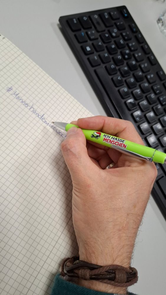Der User schreibt mit einem hellgrünen Kugelschreiber, auf dem „Ich hasse Menschen“ steht, auf kariertes Papier. Auf dem Papier ist der handgeschriebene Hashtag „#Männerhandemittwoch“ zu sehen. Im Hintergrund liegt eine schwarze Computertastatur auf einem grauen Schreibtisch.