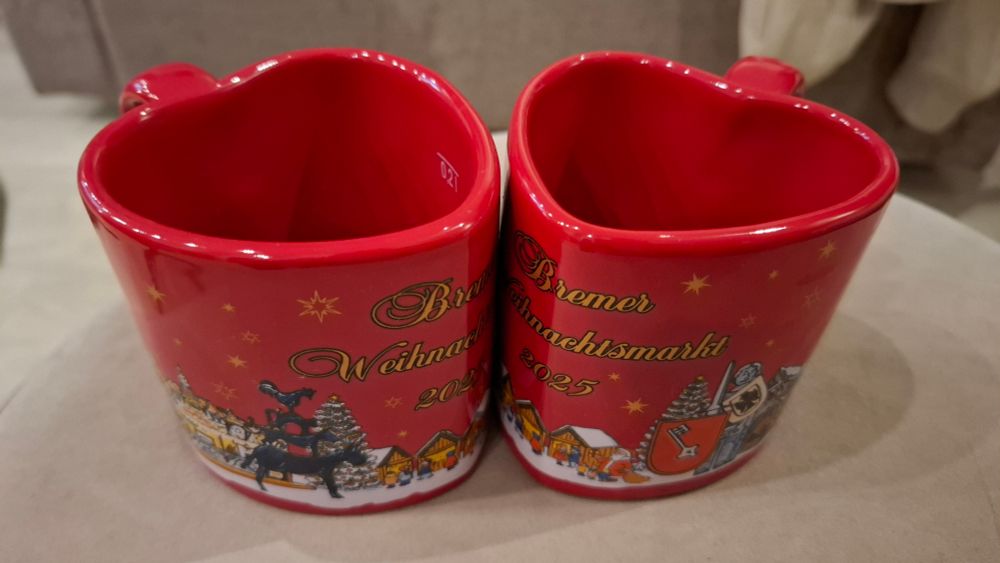 Zwei rote, herzförmige Keramikbecher stehen eng nebeneinander. Auf der Vorderseite der Becher ist in goldener Schrift „Bremer Weihnachtsmarkt 2025“ zu lesen, umgeben von kleinen Sternmotiven. Darunter ist eine winterliche Markt-Szene mit Buden, Weihnachtsbaum und u. a. den Bremer Stadtmusikanten abgebildet. Die Becher glänzen und stehen auf eine. hellen, mit weichem Stoff bezogenen Hocker. 